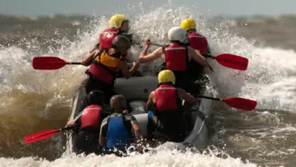 Groep mensen die branding raften op de Noordzee, peddelend in een grote raftboot terwijl ze de golven trotseren bij Wijk aan Zee. De deelnemers lachen en werken samen om de krachtige branding te overwinnen en terug te surfen naar het strand.
