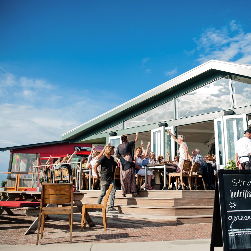 Groep medewerkers geniet van een Outstanding bedrijfsuitje in een exclusief strandpaviljoen, terwijl ze borrelen en dineren op het terras met uitzicht op zee.